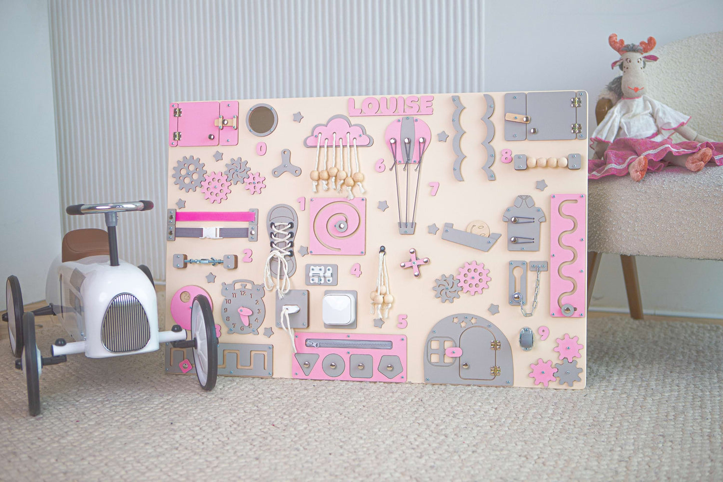 Children's Montessori busy board with pink and gray designs in a room with a toy car and plush toy.